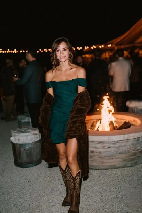 A woman in a deep green off-the-shoulder dress, slouchy brown cowboy boots, and a luxurious velvet wrap