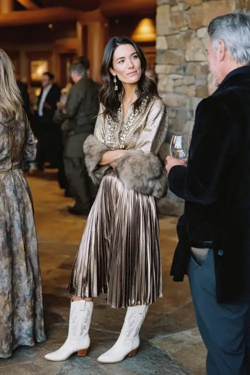 A woman in a metallic pleated skirt and embellished blouse, wearing ivory cowboy boots and a faux fur stole