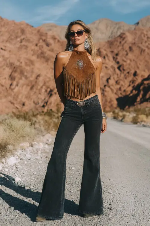 A woman in high-waisted flared jeans with a fringe suede halter top, black cowboy boots, oversized sunglasses, and silver statement earrings