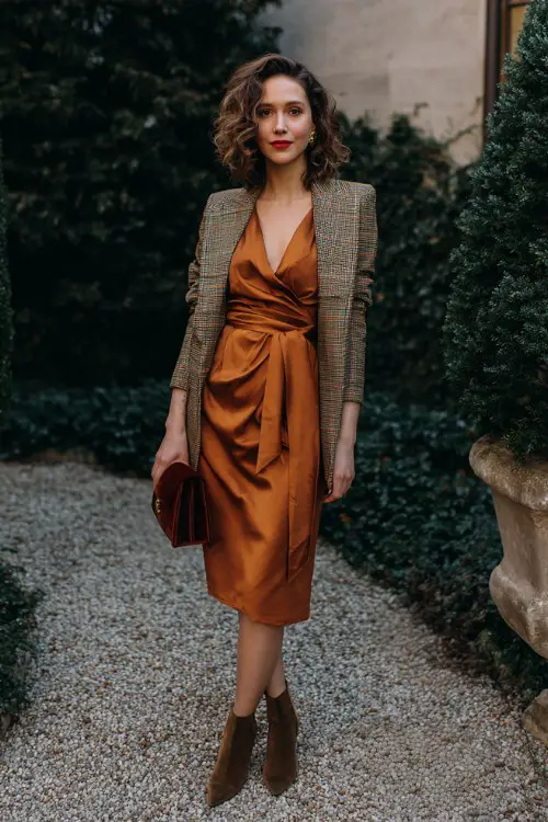 A woman over 30 wears a belted wrap dress in copper crepe with a structured plaid blazer and low suede ankle boots