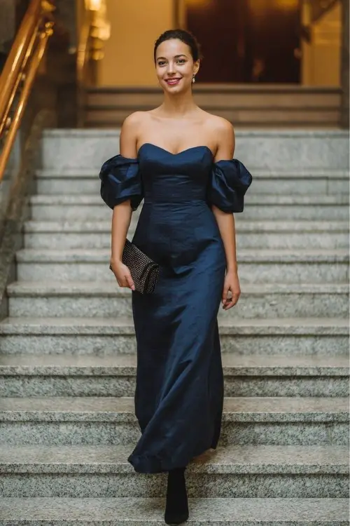 A woman over 30 wears a classic floor-length navy gown with structured shoulders, styled with stiletto-heeled black boots and a clutch