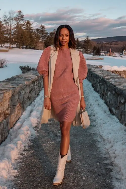 A woman over 30 wears a dusty rose midi sweater dress with puff sleeves, layered with a beige wool trench and short white cowboy boots