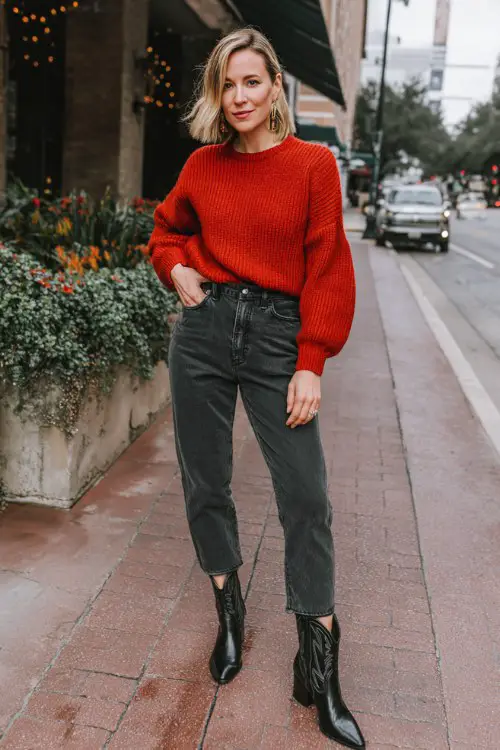 A woman over 30 wears a fitted red sweater with straight-leg dark denim and glossy black cowboy boots