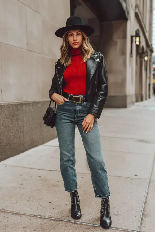 A woman over 30 wears a fitted red turtleneck sweater with a black leather jacket, high-rise jeans, black cowboy boots, and a structured black fedora
