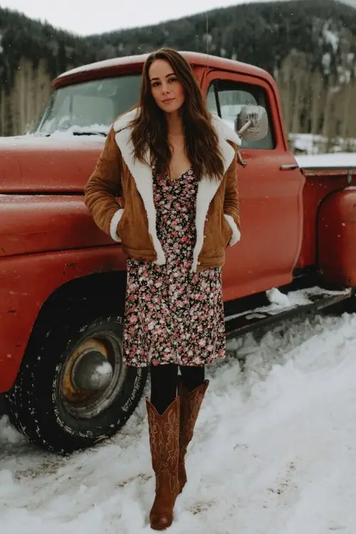 A woman over 30 wears a long-sleeve floral dress with black tights and tall brown cowboy boots, layered with a cropped teddy jacket