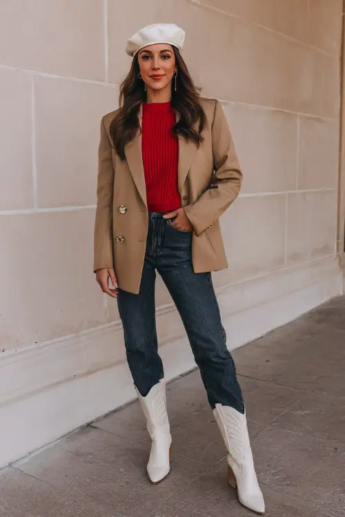 A woman over 30 wears a red crewneck sweater tucked into dark denim, topped with a tailored camel blazer, white cowboy boots, and a chic beret