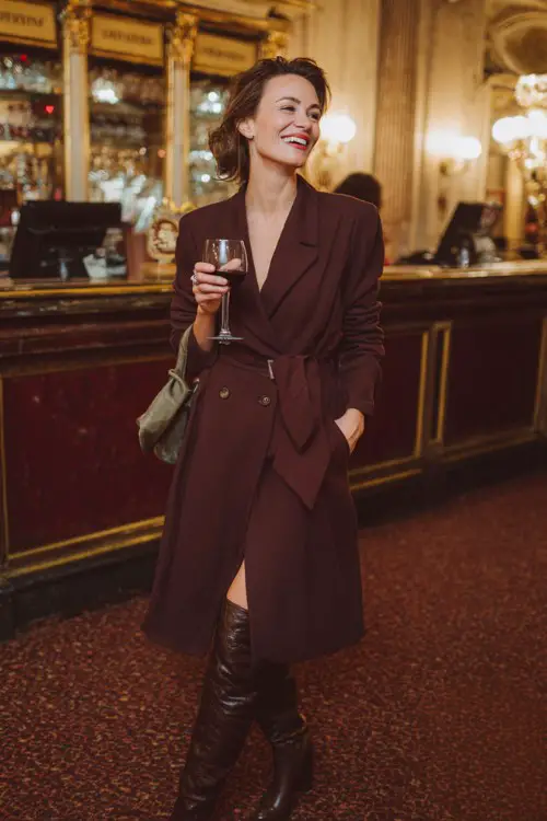 A woman over 40 wears a midi wrap dress in deep burgundy with tall leather boots and a structured wool coat