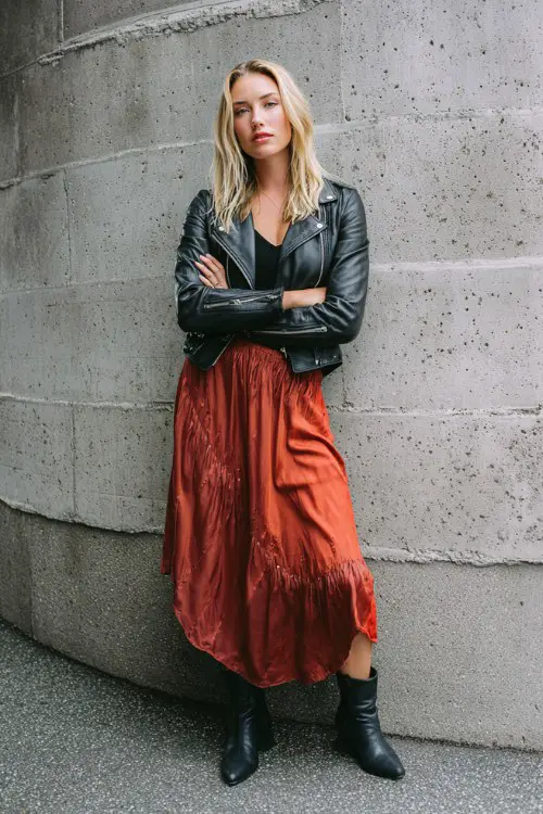 A woman wears a flowy midi skirt with a knit top and a cropped moto jacket, paired with sleek booties