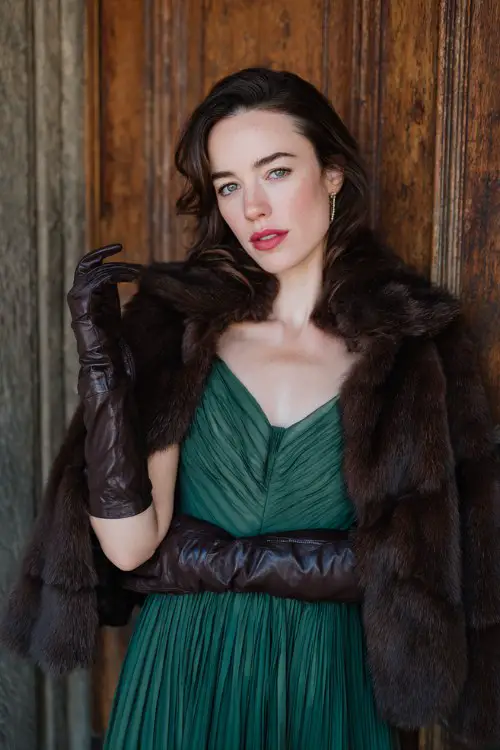 A woman wears a forest green pleated chiffon dress with a dramatic cape overlay, paired with a dark brown fur coat and leather gloves
