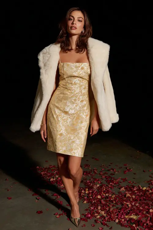 A woman wears a golden jacquard dress with a square neckline and subtle shimmer, styled with a structured cream fur collar coat and metallic heels