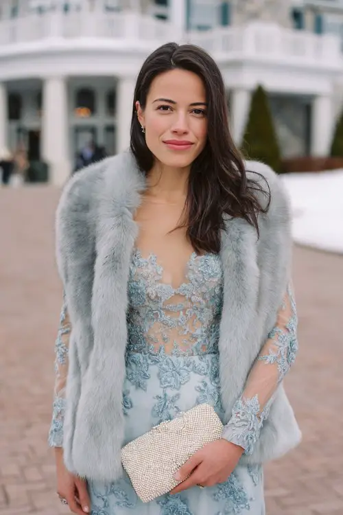 A woman wears a powder blue embroidered gown with sheer sleeves and scalloped hems, topped with a silver fox faux fur coat and pearl clutch