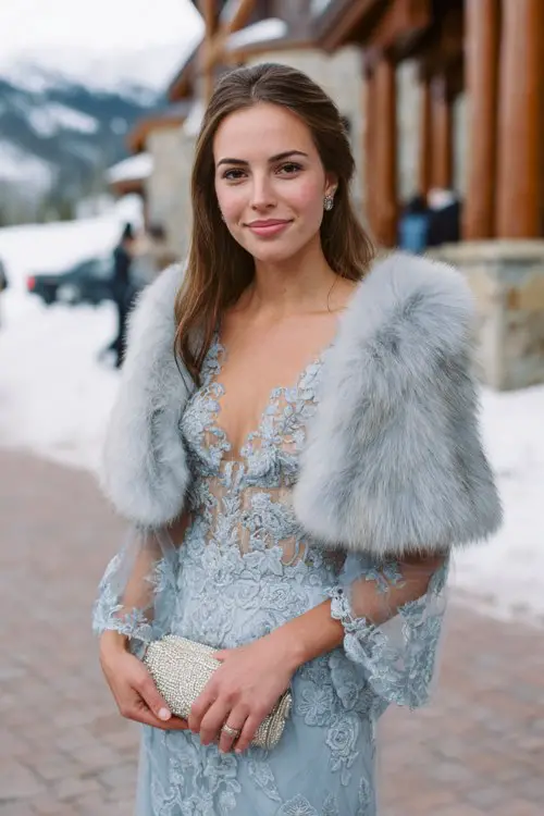 A woman wears a powder blue embroidered gown with sheer sleeves and scalloped hems, topped with a silver fox faux fur stole and pearl clutch