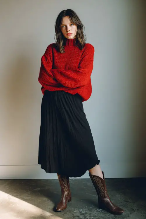 A woman wears a slouchy red sweater tucked slightly into a black pleated midi skirt with brown western boots