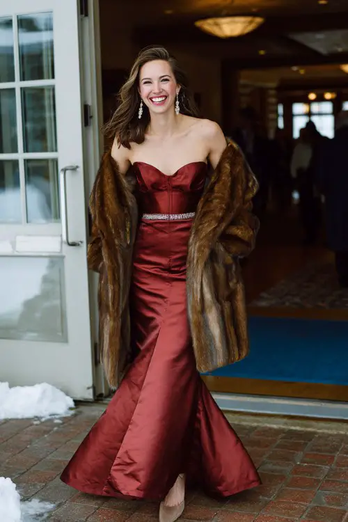 A woman wears a wine-red satin mermaid gown with a structured bodice and pearl belt with a long fur coat, gold drop earrings, and matching pumps