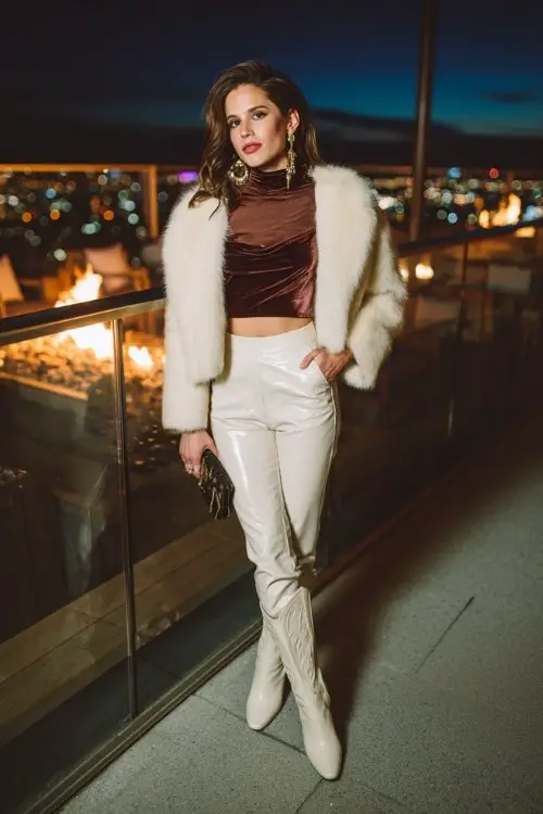 A woman wears leather-look leggings with white cowboy boots, a cropped faux-fur jacket layered over a slim turtleneck and satin camisole, styled with bold clutch and hoop earrings