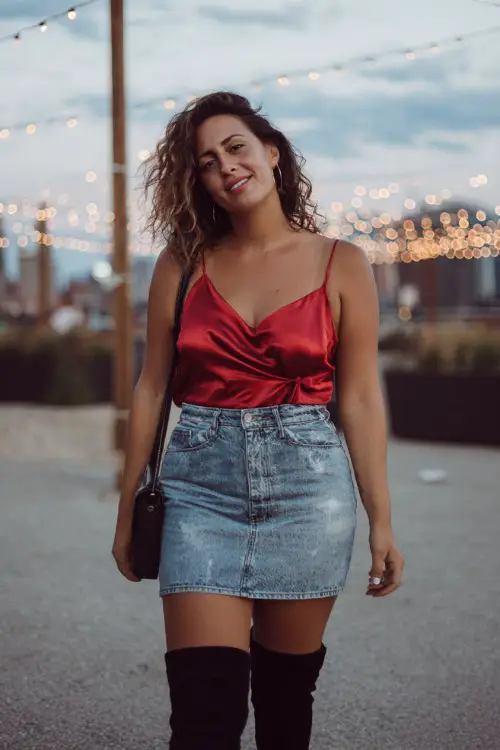 A curvy woman over 30 wears a silky red cami top with a denim mini skirt, black thigh-high boots, and a crossbody bag
