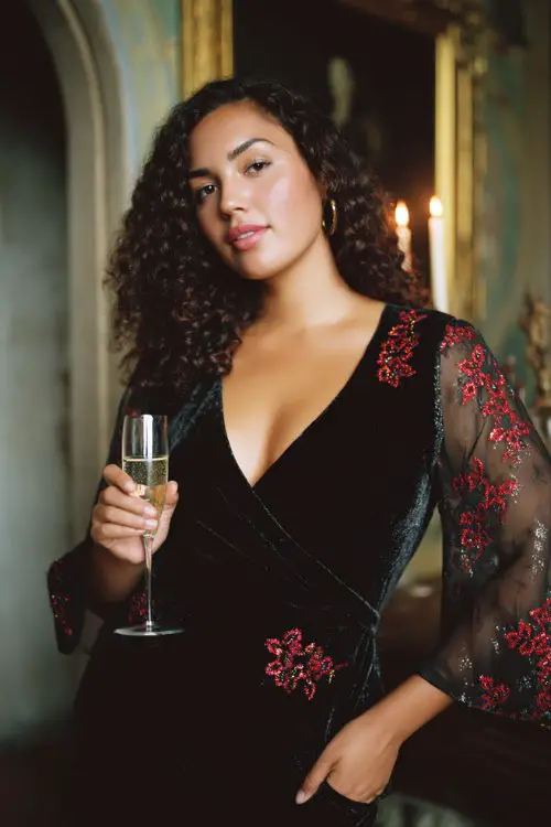 A curvy woman wears a fitted black velvet wrap dress with red sequin floral embroidery and sheer bell sleeves