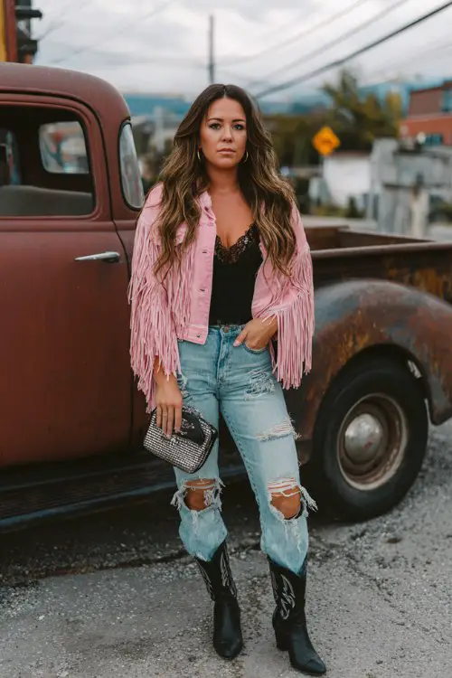 A curvy woman wears a pink fringe jacket over a lace top and distressed jeans, black cowboy boots