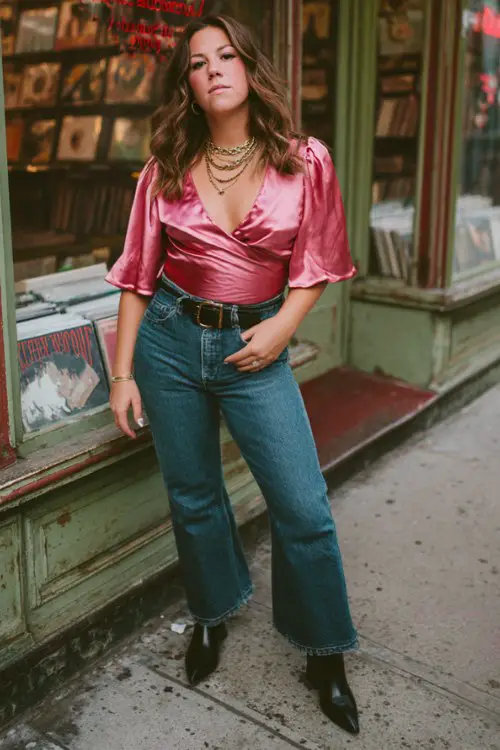 A curvy woman wears a pink satin wrap top with flared jeans, black cowboy boots, and layered necklaces
