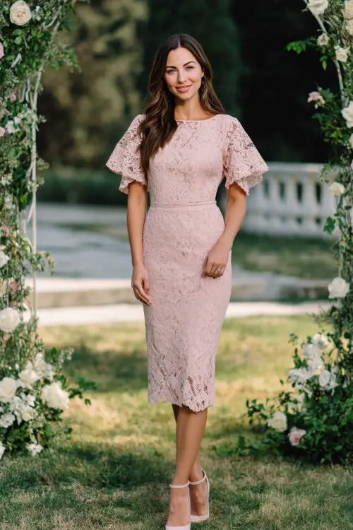 A curvy woman wears a romantic lace midi formal dress in blush pink with flutter sleeves and defined waist, styled with elegant block heels