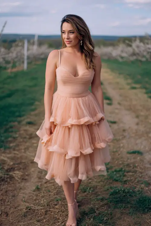 A curvy woman wears a soft peach tulle midi dress with ruffled skirt layers and fitted waist, styled with elegant heeled sandals