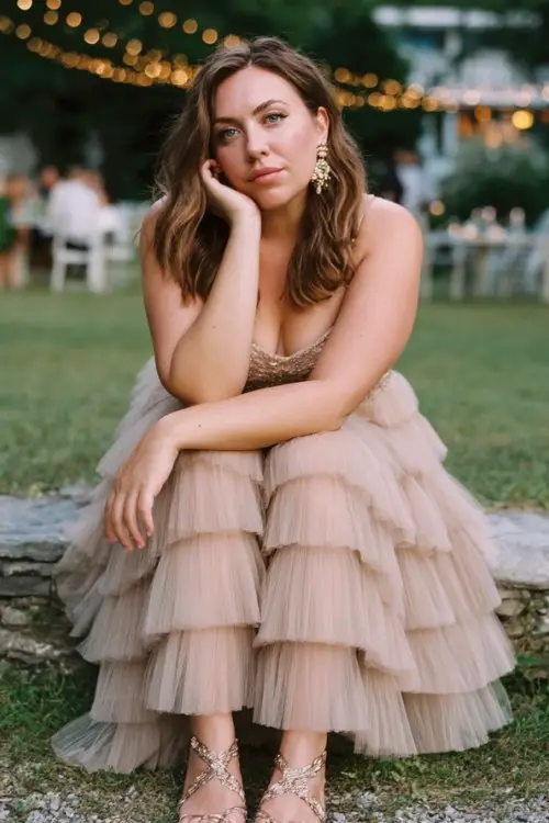 A curvy woman wears a tiered tulle dress with subtle shimmer details, delicate sandals, and statement earrings, outdoor garden reception with fairy lights