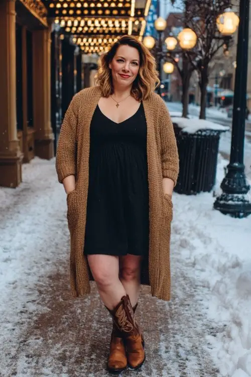 A plus size woman over 40 wears a long camel cardigan over a fitted black dress with brown cowboy boots