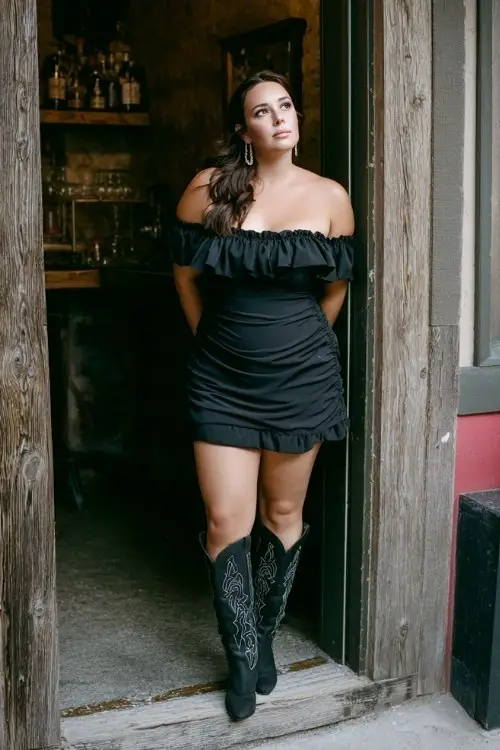 A plus-size woman wears a black off-shoulder mini dress with ruffle detail and tall black cowboy boots