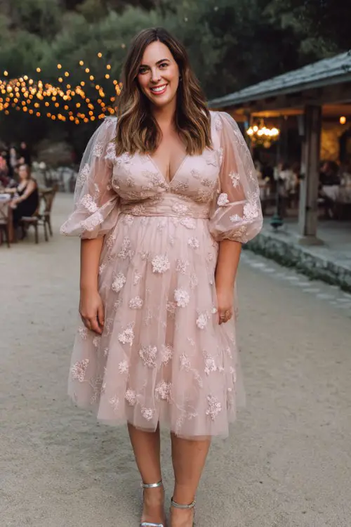 A plus-size woman wears a blush pink tulle midi dress with embroidered florals and sheer puff sleeves, paired with silver ankle-strap heels