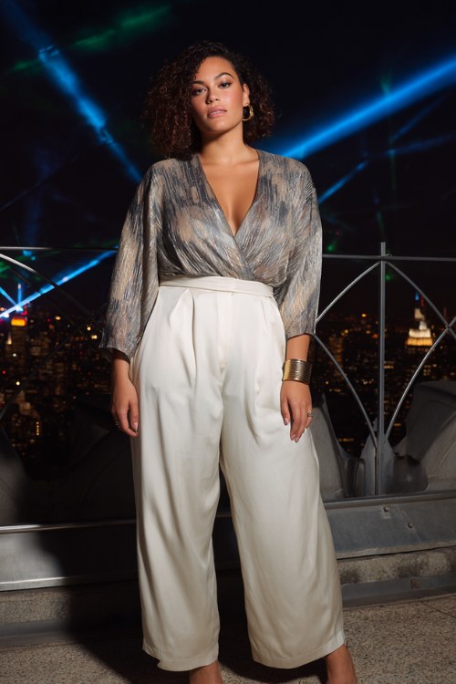 A plus-size woman wears a liquid-metal draped top with wide-leg satin pants, paired with sleek heels and bold cuffs