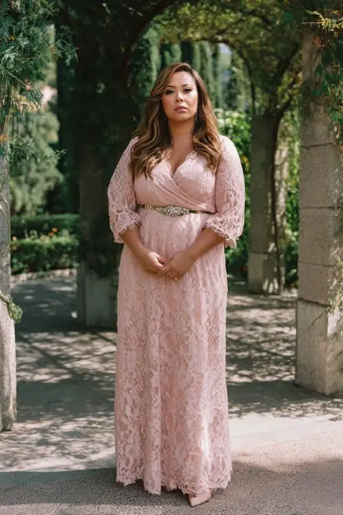 A plus-size woman wears a romantic blush lace maxi dress with sheer sleeves, classic pumps, and a jeweled belt, garden arch ceremony background