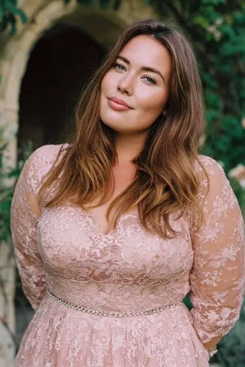 A plus-size woman wears a romantic blush lace maxi dress with sheer sleeves, classic pumps, and a jeweled belt