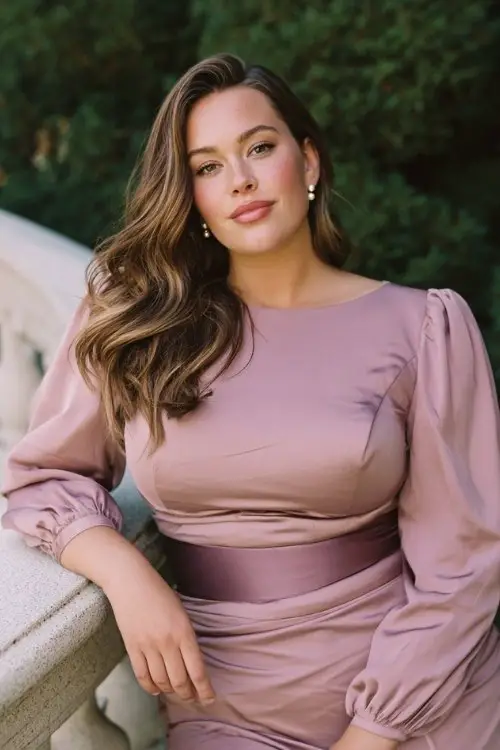 A plus-size woman wears a sculpted midi dress with subtle pleats and satin belt, closed-toe heels, and pearl drop earrings