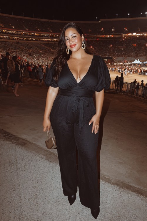 A plus-size woman wears a sleek black jumpsuit with a dramatic neckline and a cinched waist, styled with pointed-toe boots and bold earrings