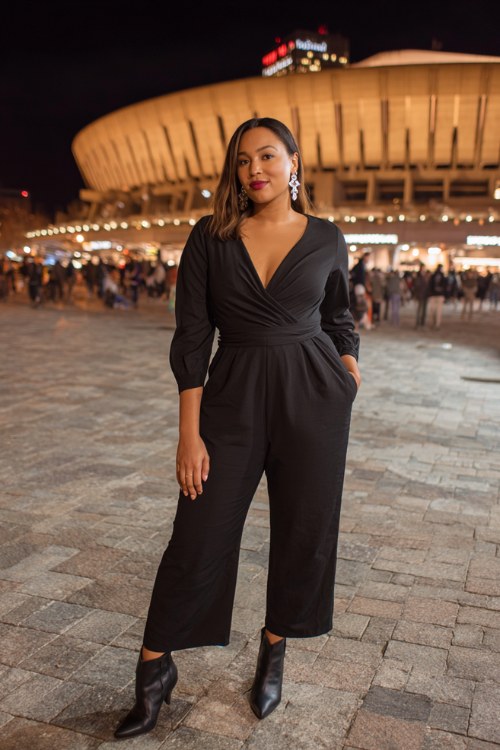 A plus-size woman wears a sleek black jumpsuit with a dramatic neckline and a cinched waist, styled with pointed-toe boots