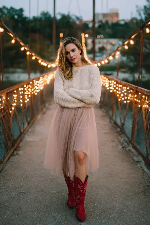 A woman on a bridge wrapped in warm glowing string lights, wearing a cream knit sweater tucked into a flowing blush pink tulle skirt and red high-heeled cowboy boots
