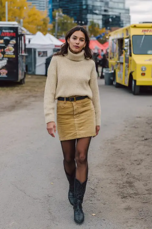 A woman over 30 wears a corduroy mini skirt with black tights, a chunky turtleneck sweater, and classic cowboy boots, walking past food trucks