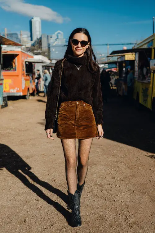 A woman over 30 wears a corduroy mini skirt with black tights, a chunky turtleneck sweater, and classic cowboy boots