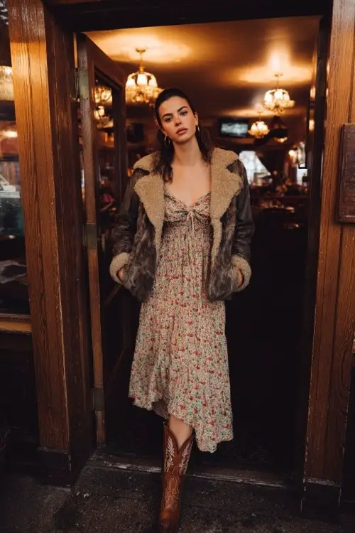 A woman over 30 wears a floral midi dress layered with a cropped faux fur jacket and brown embroidered cowboy boots