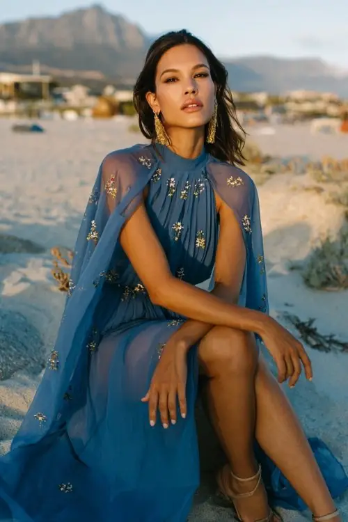 A woman over 30 wears a flowing blue chiffon maxi dress with a sheer cape overlay embroidered with tiny blossoms, strappy heels, and gold drop earrings, wedding setting at sunset