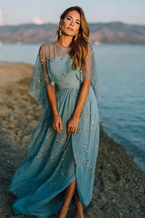 A woman over 30 wears a flowing blue chiffon maxi dress with a sheer cape overlay embroidered with tiny blossoms, strappy heels