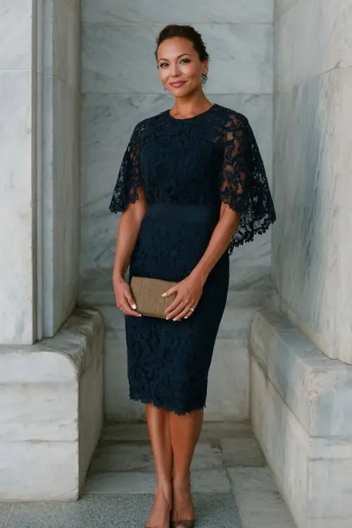 A woman over 40 wears a lace-overlay navy sheath dress with delicate cape detail, polished pumps, and a clutch bag, historic chapel wedding setting