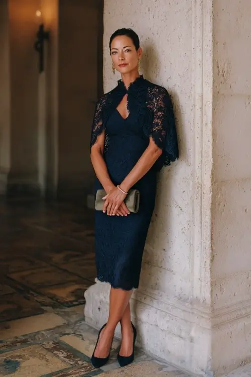 A woman over 40 wears a lace-overlay navy sheath dress with delicate cape detail, polished pumps, and a clutch bag