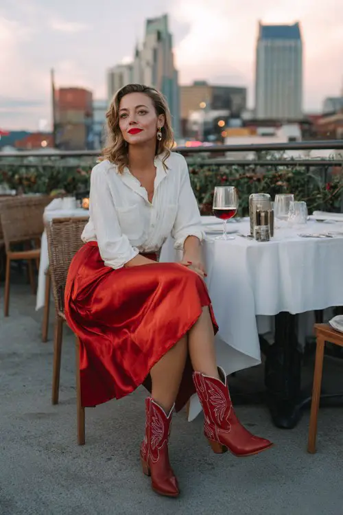 A woman over 40 wears a midi red satin skirt with a white tucked-in blouse, bold lipstick, and red cowboy boots