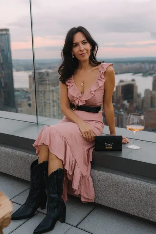 A woman over 40 wears a pink ruffle-hem midi dress with a belted waist and sleek black cowboy boots