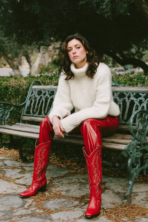 A woman sitting pensively on a vintage iron park bench, wearing a chunky white knit sweater and fitted red leather pants