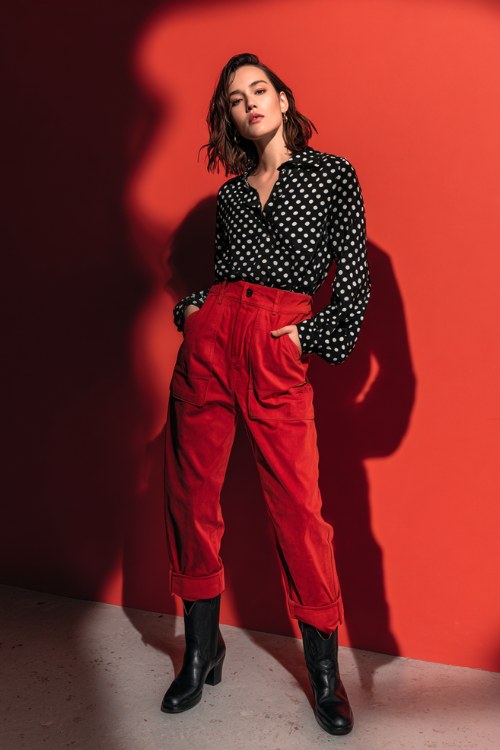 A woman wears a black-and-white polka dot blouse tucked into red high-waisted trousers, paired with black cowboy boots