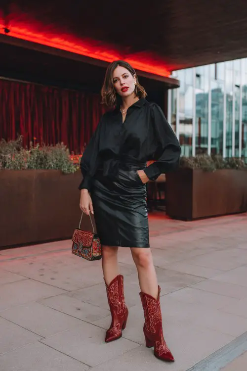 A woman wears a black faux leather pencil skirt with a tucked-in silky blouse and red embroidered cowboy boots