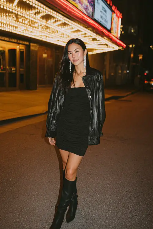 A woman wears a black fitted mini dress with a structured leather jacket draped over her shoulders, paired with knee-high boots and bold silver earrings