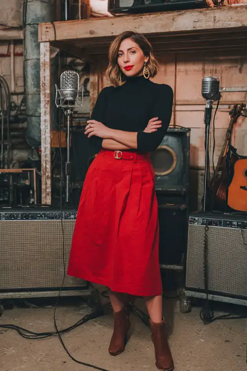 A woman wears a black fitted turtleneck and a red pleated midi skirt with ankle boots